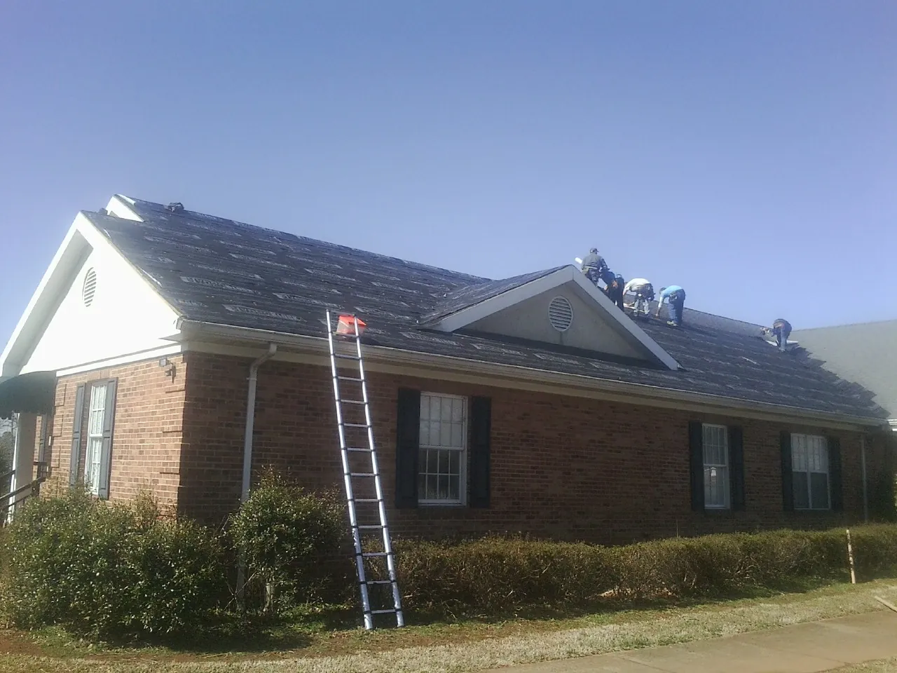 Crew tearing shingles off a brick ranch roof with a ladder set — Emergency Roof Repair work by our crew in Deming