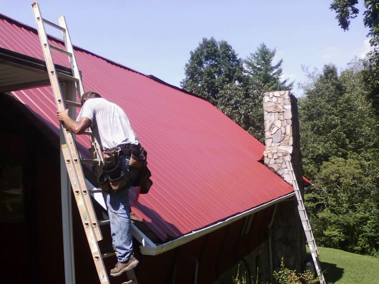 Roofer on a ladder beside a red metal roof and stone chimney — Soffit & Fascia Repair by Roofing Brothers in Deming